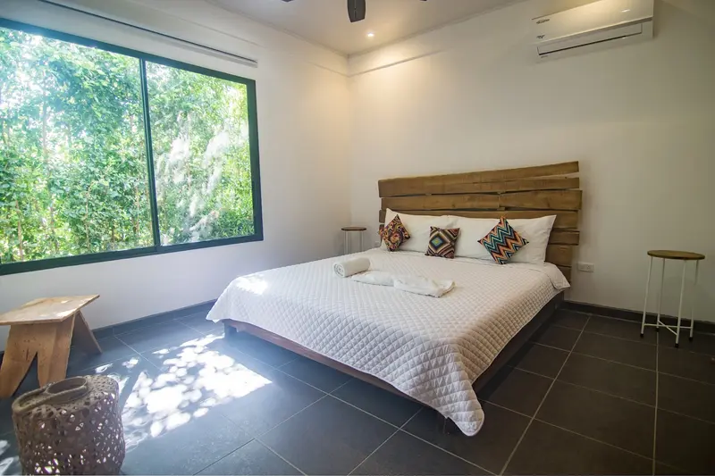 Dormitorio con cama grande de cabecera de madera, ventanal al jardín y aire acondicionado