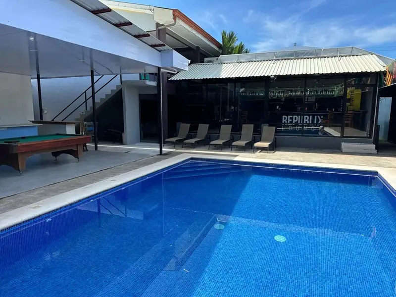 Piscina vista desde el borde, con una mesa de billar a la izquierda y tumbonas bajo el edificio del fondo