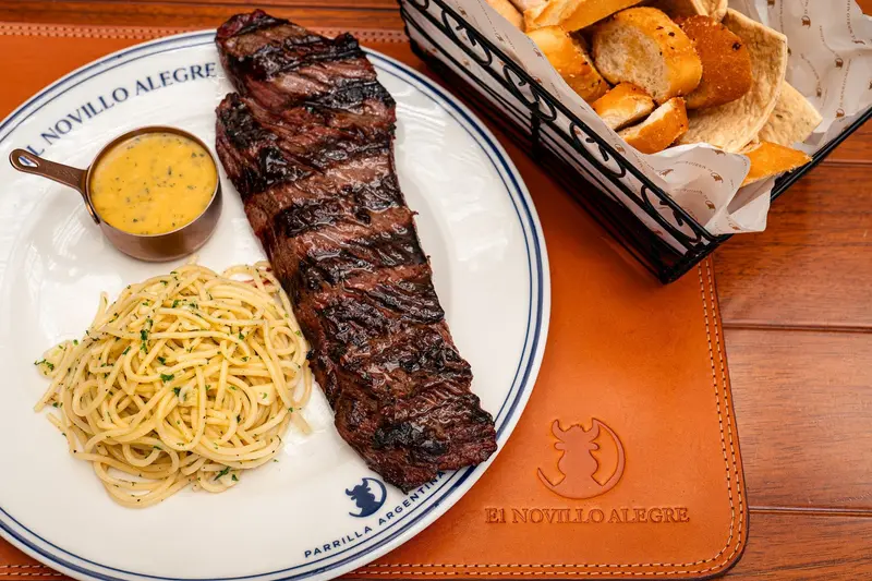 Corte de carne a la parrilla con spaghetti, una salsa amarilla en cazuelita y una canasta de pan