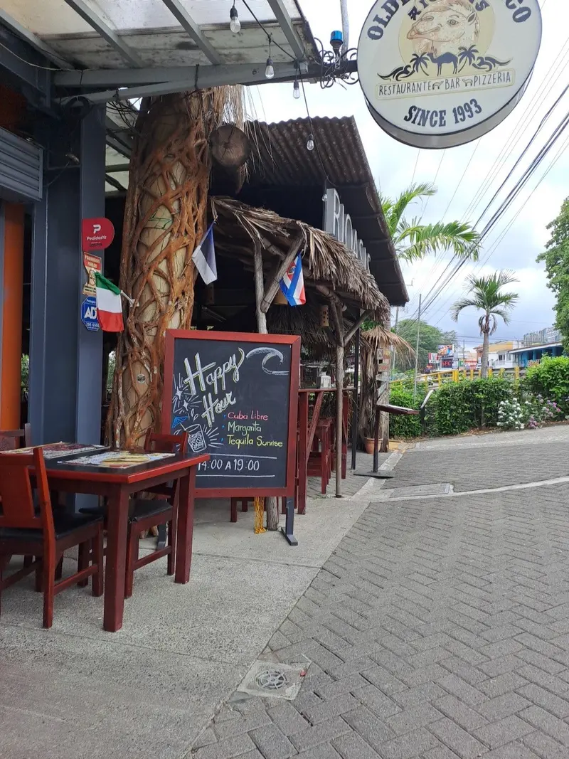 Frente del restaurante con terraza, una mesa de madera, una pizarra y el letrero redondo «Since 1993»
