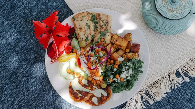 Plato con pan, garbanzos, hojas verdes y salsas sobre un mantel azul, con una flor de hibisco