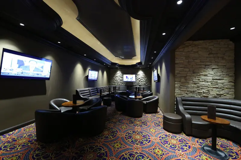 Salón del casino con sillones de cuero negro, alfombra estampada y pantallas en la pared de piedra