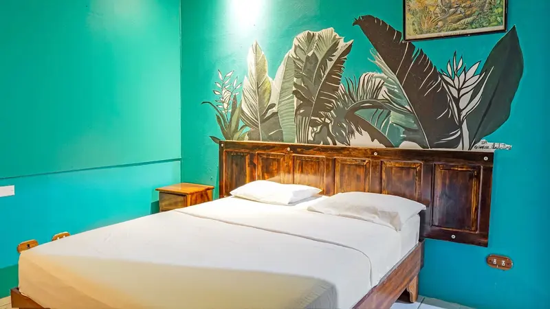 Habitación con cama doble, cabecera de madera y pared turquesa pintada con hojas tropicales