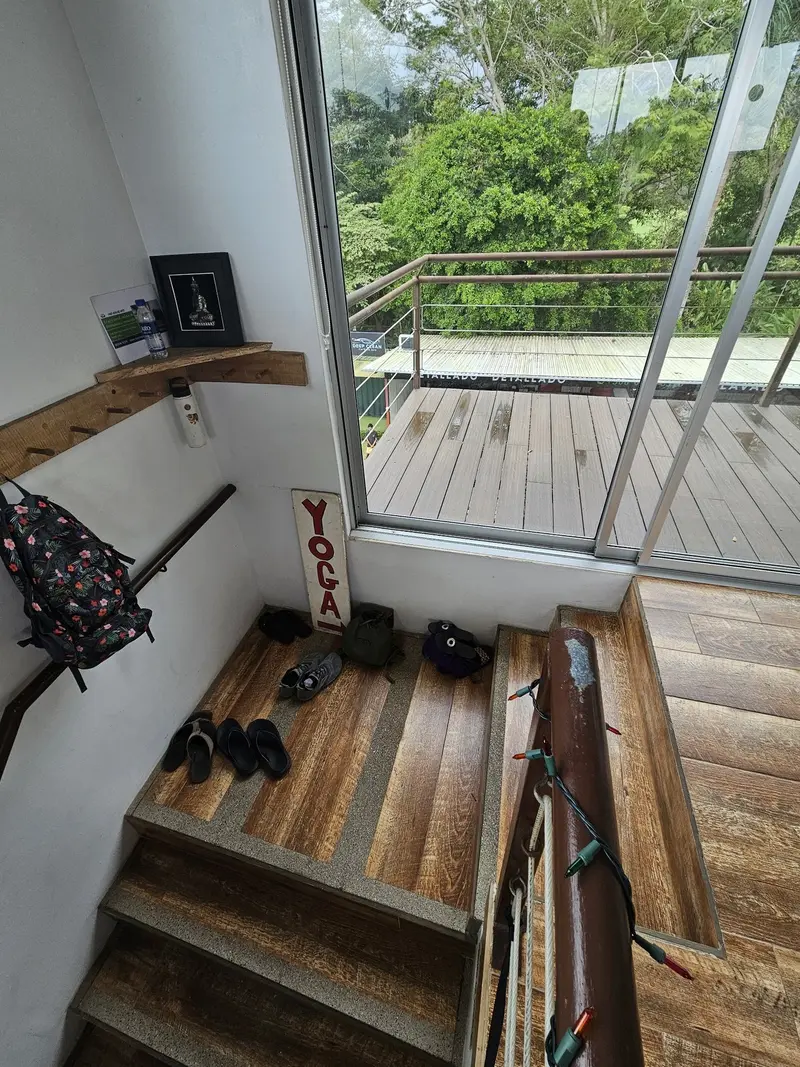 Entrada del estudio con zapatos junto a la escalera de madera, un letrero de yoga y vista a la selva