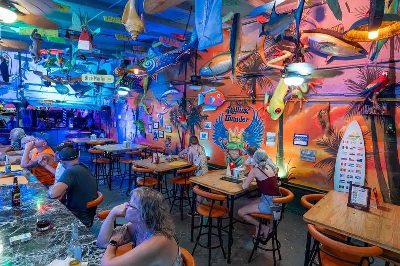 Salón con paredes pintadas de colores, mesas de madera y banquetas altas bajo un techo decorado