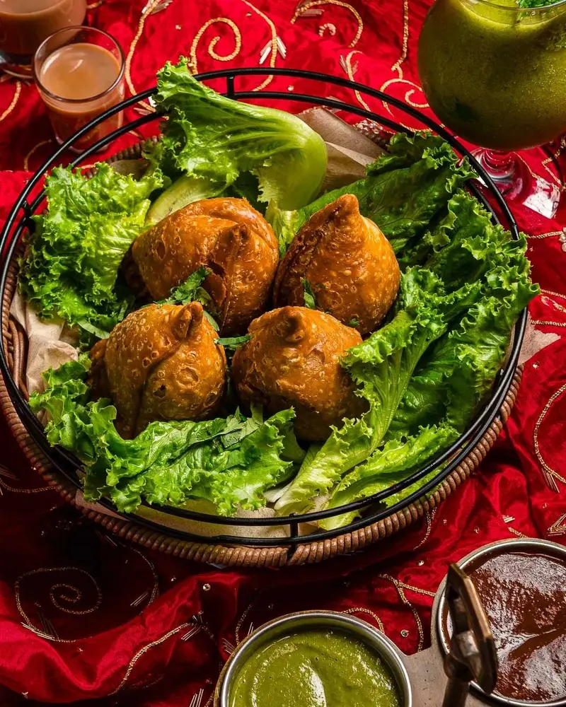 Samosas de papa sobre hojas de lechuga en un plato de metal, con chutneys verde y de tamarindo alrededor