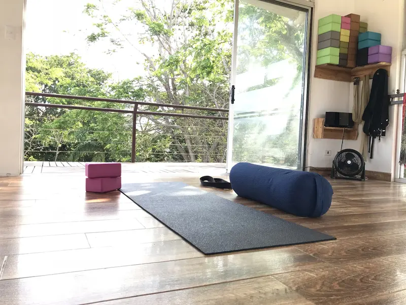 Esterilla gris con un bolster azul y bloques de yoga en la terraza del estudio, con vista a los árboles