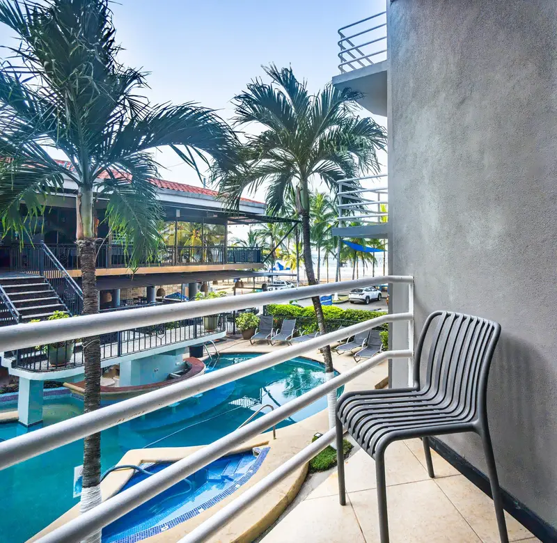 Balcón con una silla gris sobre la piscina, las palmeras y el mar al fondo