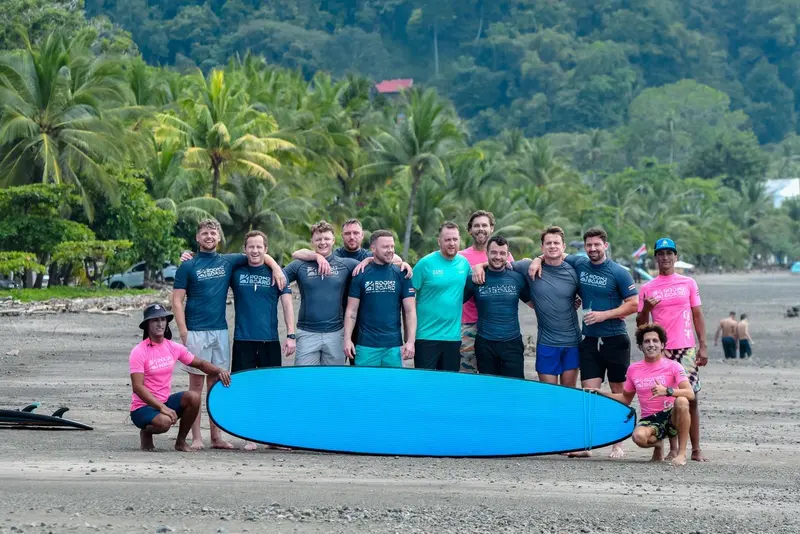 Grupo con licras azules y una tabla azul grande sobre la arena tras una clase de surf