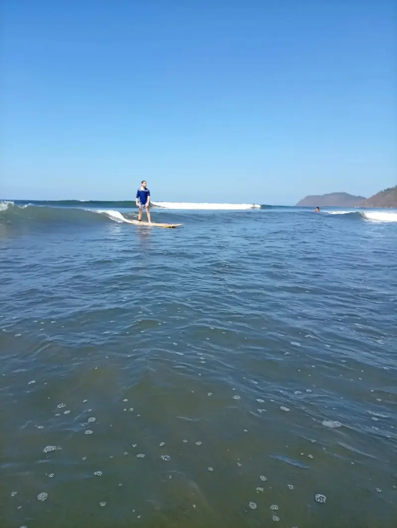 Surfista lejano sobre una ola en aguas azules, con la costa al fondo