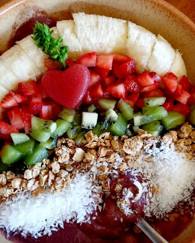 Bowl de açaí visto desde arriba con fresa, kiwi, banano, granola y coco rallado, con un corazón de sandía