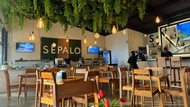 Salón con mesas y sillas de madera, plantas colgantes en el techo y el rótulo Sêpalo en la pared del fondo