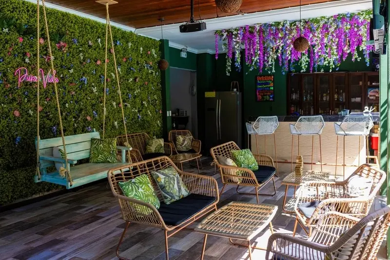 Lounge con columpio, sillones de ratán, barra y pared verde con flores