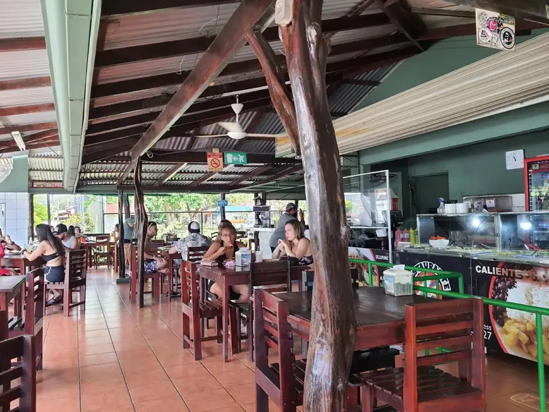 Salón con mesas y sillas de madera bajo un techo de vigas, con la barra del buffet al fondo
