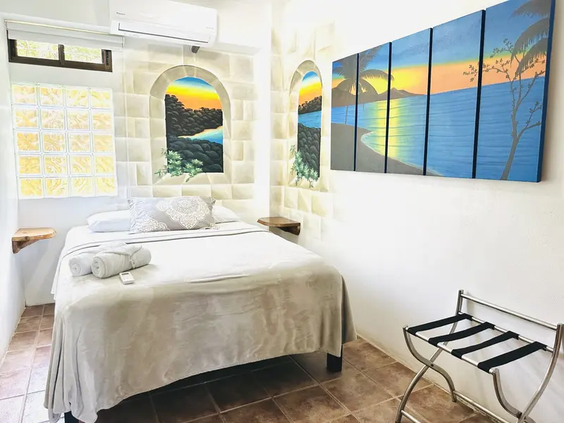 Habitación con cama blanca, luminarias, equipaje sobre un soporte y un cuadro de atardecer