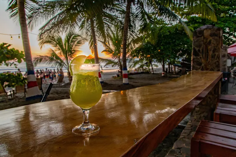 Vaso con una bebida verde sobre la barra de madera, con palmeras y la playa al fondo
