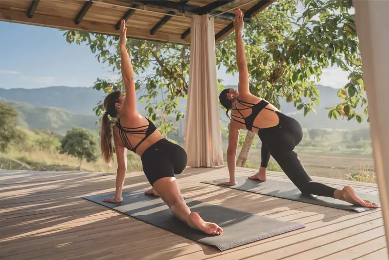 Dos personas hacen yoga sobre tapetes en una terraza de madera abierta con vista a las montañas