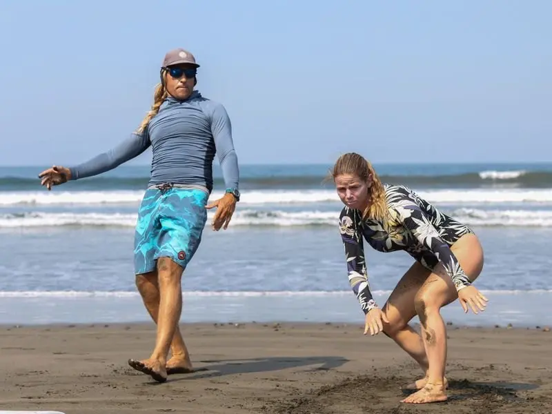 Dos personas practican la postura de surf sobre la arena, con el mar al fondo