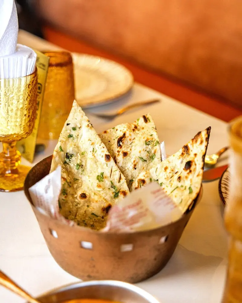 Cesta de cobre con pan naan de ajo recién horneado sobre una mesa con copas ámbar