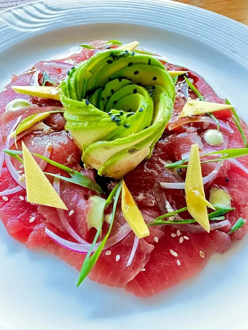 Carpaccio de atún con rosa de aguacate, cebolla morada y brotes sobre un plato blanco