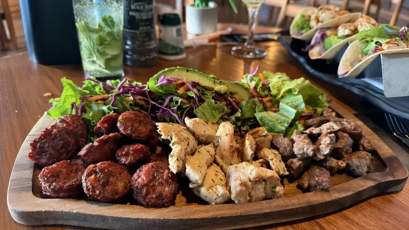 Tabla de madera con albóndigas, trozos de pollo y carne sobre ensalada, junto a un mojito y tacos