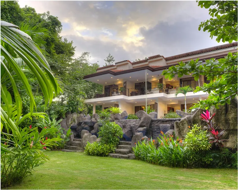 Edificio del hotel entre jardines tropicales y un muro de piedra, bajo un cielo con nubes