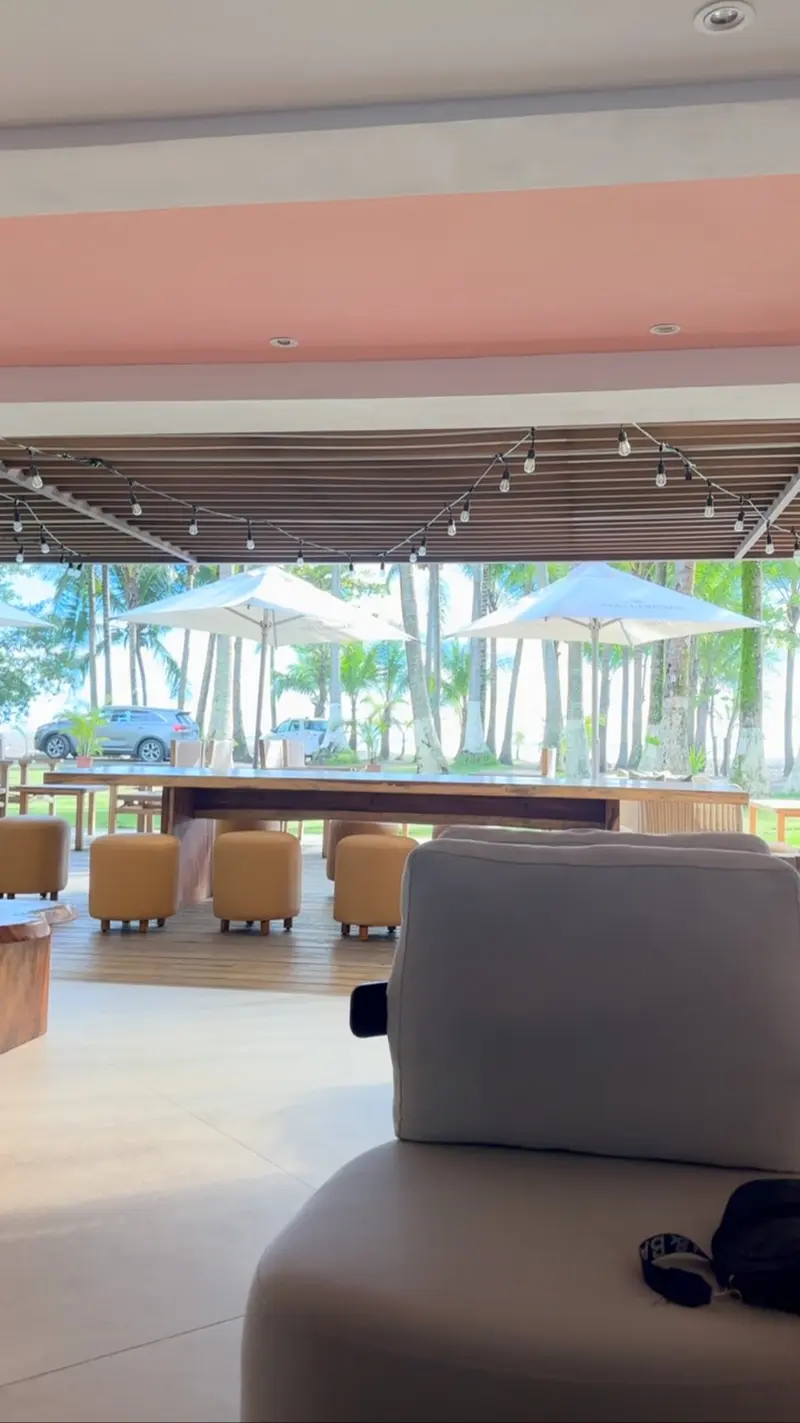 Salón abierto del restaurante con sofás y mesas, y palmeras y sombrillas al fondo