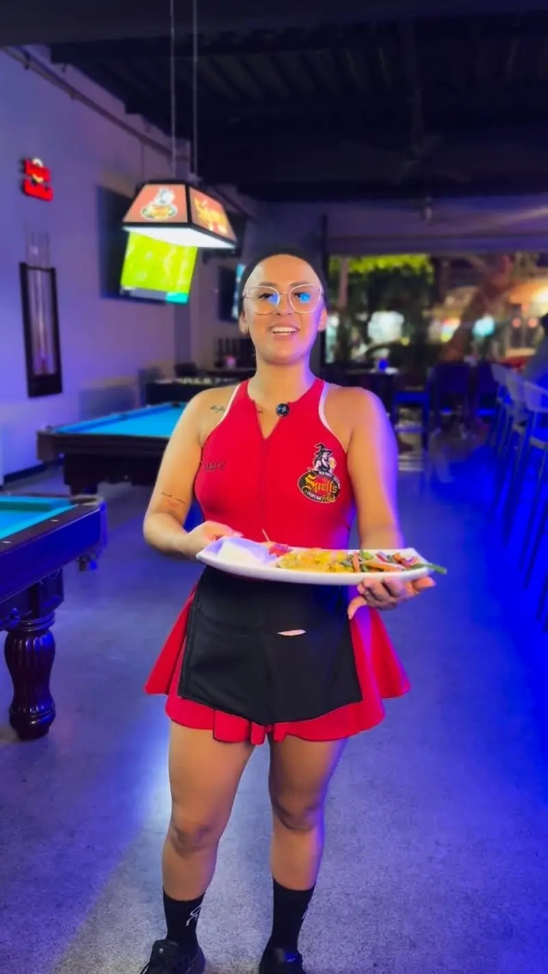Mesera con uniforme rojo y negro que lleva un plato, junto a una mesa de billar bajo luz azul