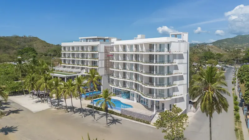 Vista aérea del edificio blanco del hotel frente a la playa, con la piscina al pie y la costa al fondo