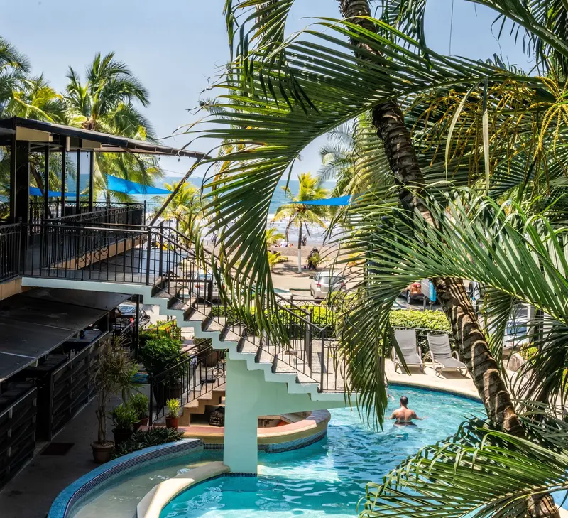 Piscina con puente de escalera y palmeras, con la playa y sombrillas azules al fondo