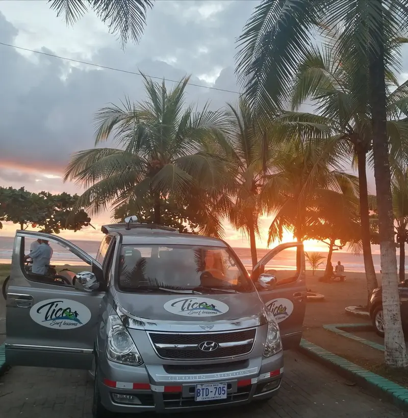 Furgoneta con el logo de Tico Surf Lessons estacionada frente a palmeras y un atardecer anaranjado
