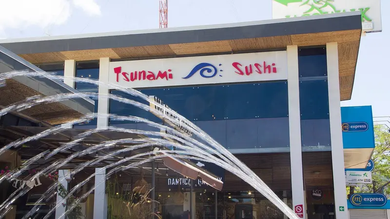 Fachada de Tsunami Sushi con su rótulo rojo y azul frente a una fuente de agua