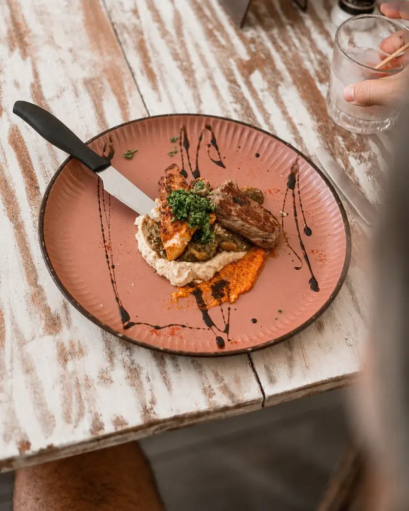 Plato rosado visto desde arriba con carne sobre puré, salsa naranja y gotas de salsa oscura, con un cuchillo al lado