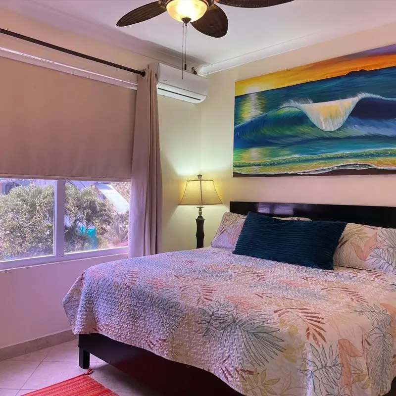 Dormitorio principal con cama, ventilador de techo, una pintura de una ola y ventana