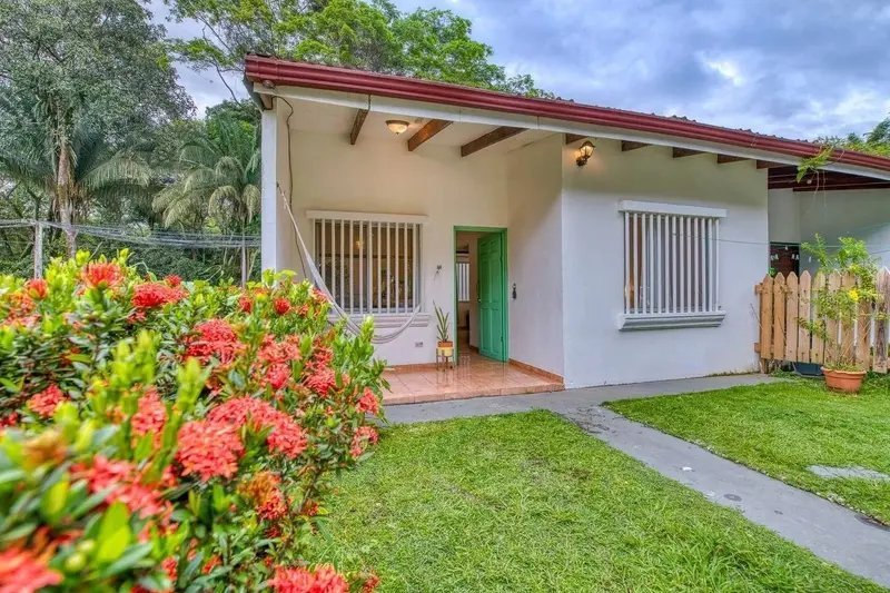 Casita blanca de techo rojo con ventanas enrejadas, rodeada de césped y flores rojas
