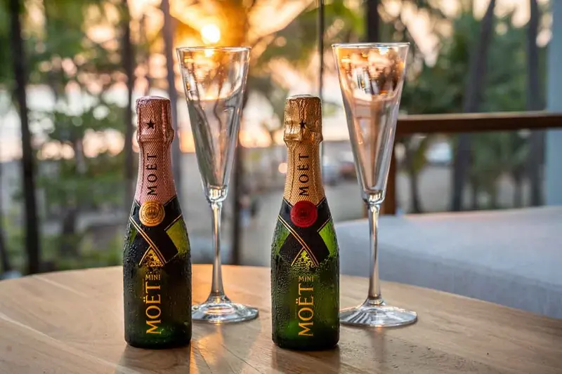 Dos botellas pequeñas de Moët y dos copas sobre una mesa, con luz de atardecer entre las palmeras