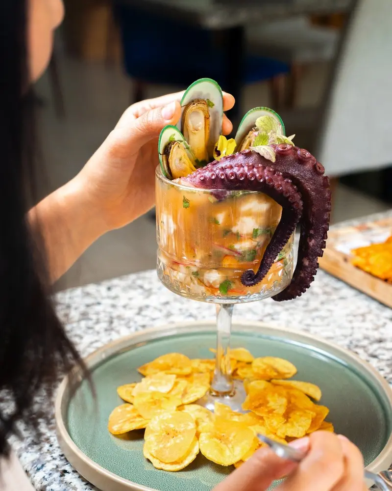 Copa de ceviche coronada con pulpo y mejillones, sobre un plato con chips de plátano