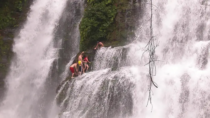 Cascada alta de caída doble en la selva, con personas pequeñas sobre la roca a media altura