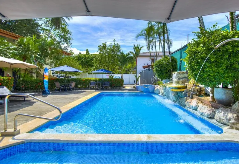 Piscina azul con camastros y sombrillas, rodeada de palmeras y un jardín