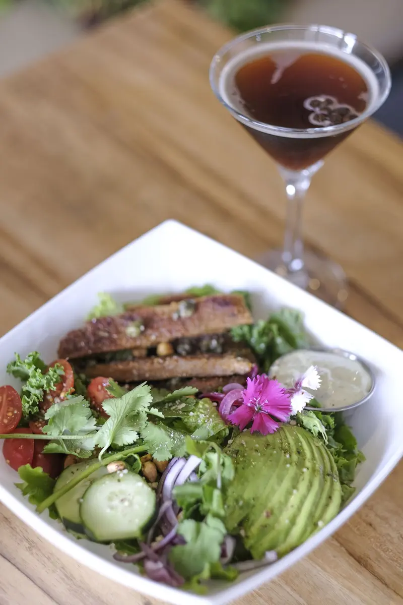 Ensalada con aguacate, pepino, tomate cherry, cilantro y rebanadas empanizadas en un plato blanco, con una copa de cóctel oscuro al fondo