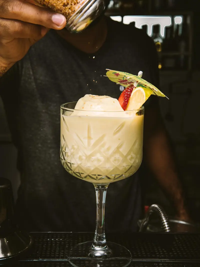 Cóctel cremoso en copa alta con una sombrilla de papel y una fresa, servido por un barman