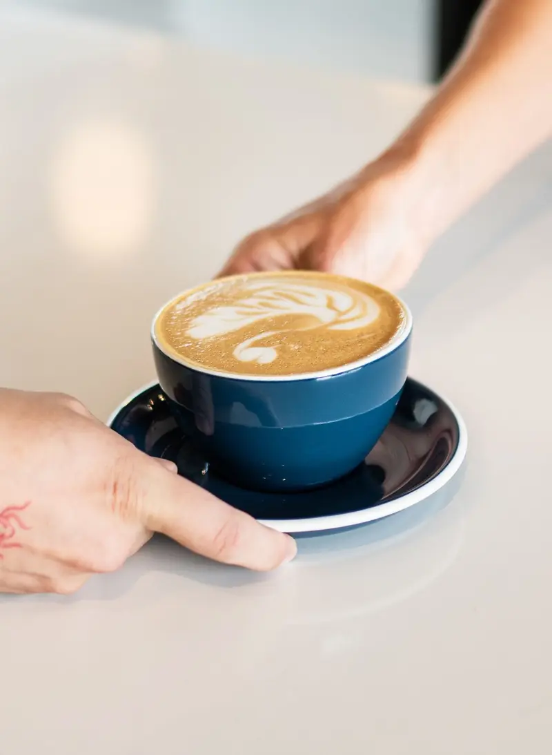Manos acercando una taza azul de café con arte latte sobre una mesa blanca