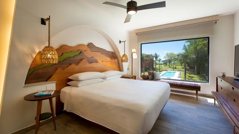 Dormitorio con cama king, cabecera de madera tallada y una ventana grande con vista a la piscina y los jardines