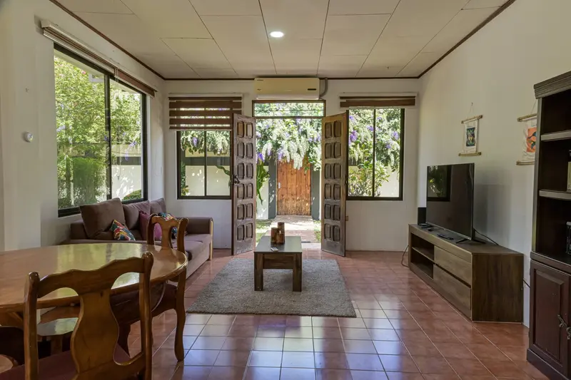 Sala de una casita con mesa de comedor de madera, sofá, televisor y puertas abiertas hacia el jardín