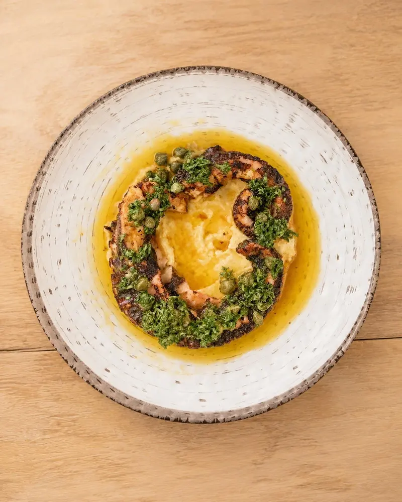 Pulpo con salsa de hierbas sobre crema amarilla en un plato blanco de borde texturizado, visto desde arriba
