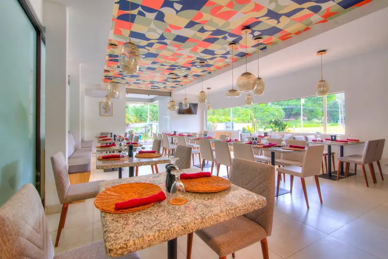 Comedor del restaurante con mesas de granito, sillas y techo de colores, con vista al jardín