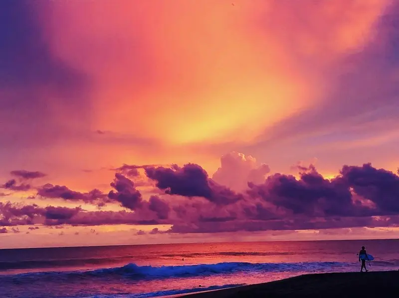 Atardecer de nubes rosadas y moradas sobre el mar, con un surfista en la orilla