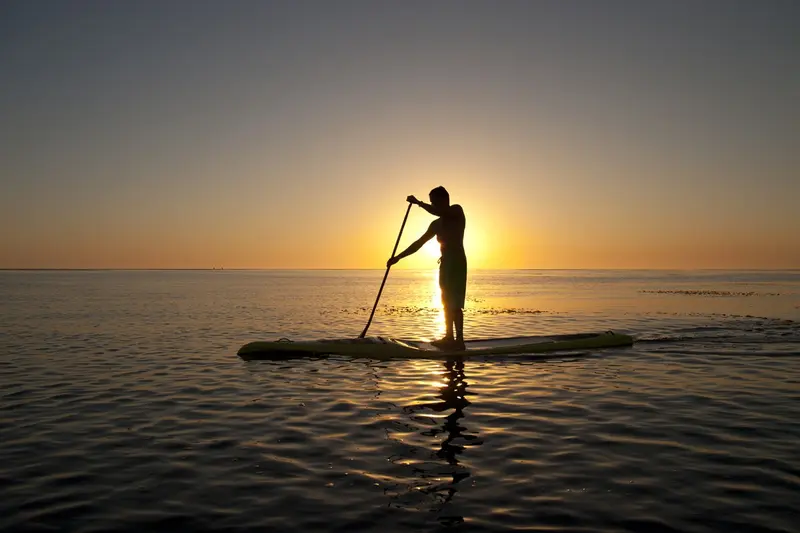 Persona de pie sobre una tabla de SUP al atardecer, a contraluz sobre el agua