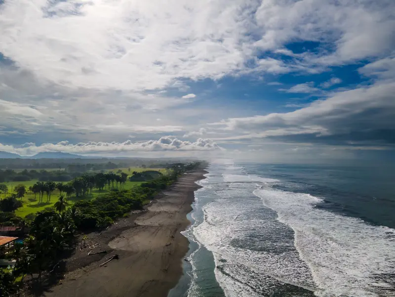 Vista aérea de la playa de arena oscura con olas y nubes sobre el mar