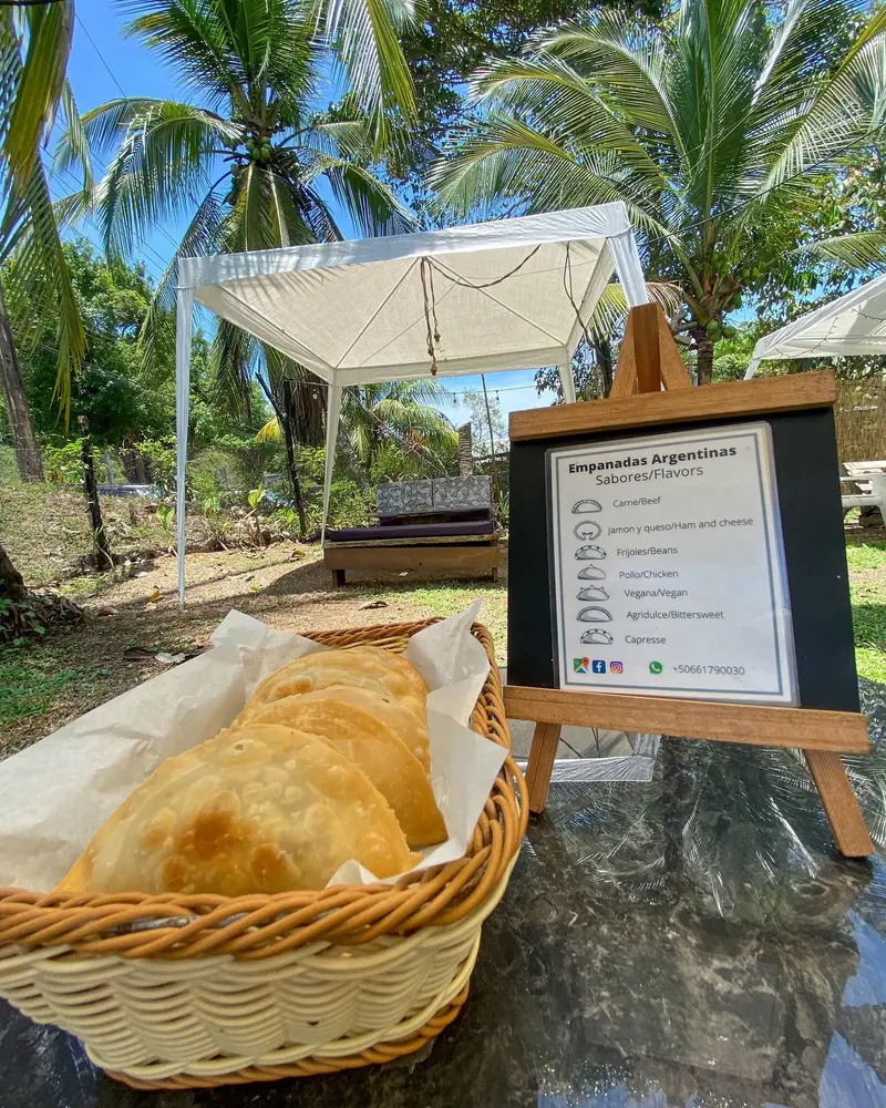 Canasta con empanadas sobre una mesa al aire libre, con un pizarrón de menú y palmeras al fondo